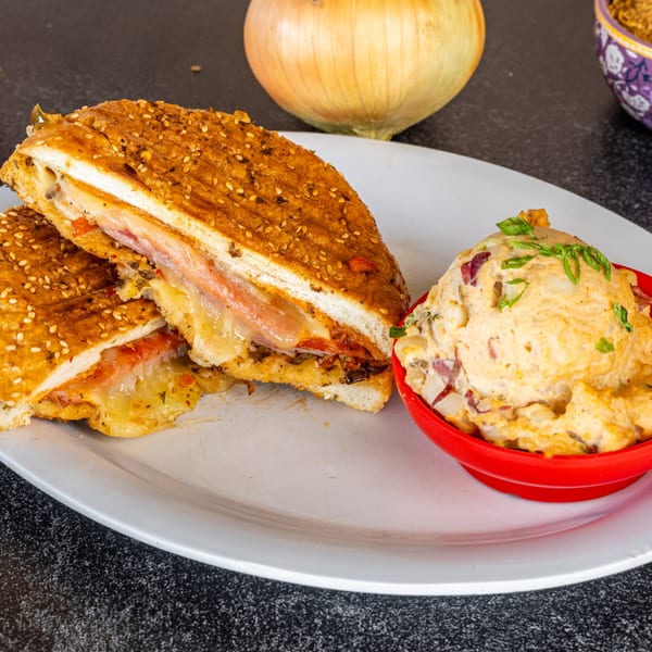 Grilled sandwich with melted cheese and meat, served with a side of potato salad in a red bowl.