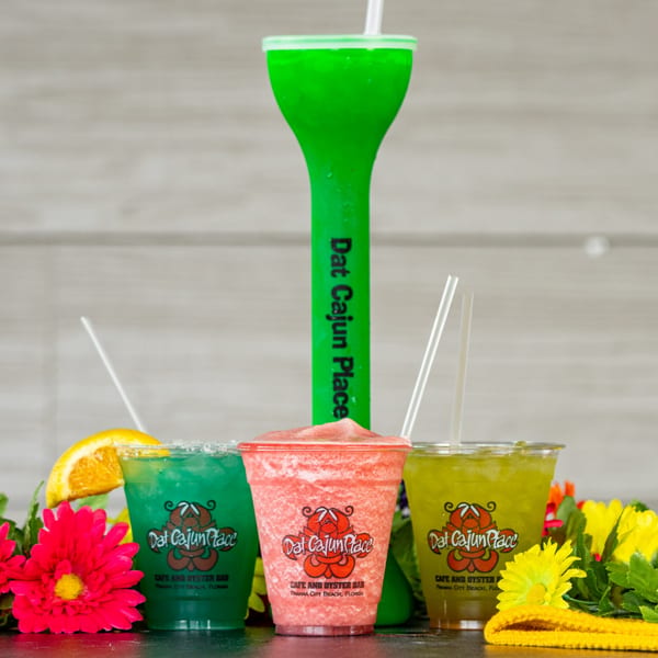 Three colorful Dat Cajun Place Cafe drinks, including a tall green one, surrounded by flowers.